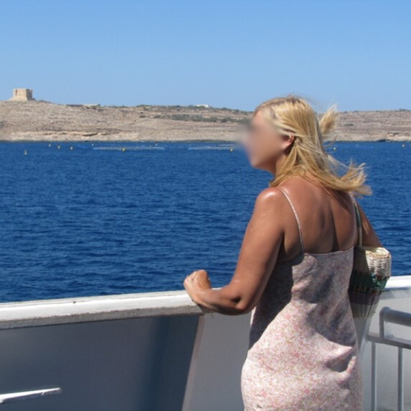 Photo of a woman with her face blurred, standing on a boat and looking at the very blue sea and sky